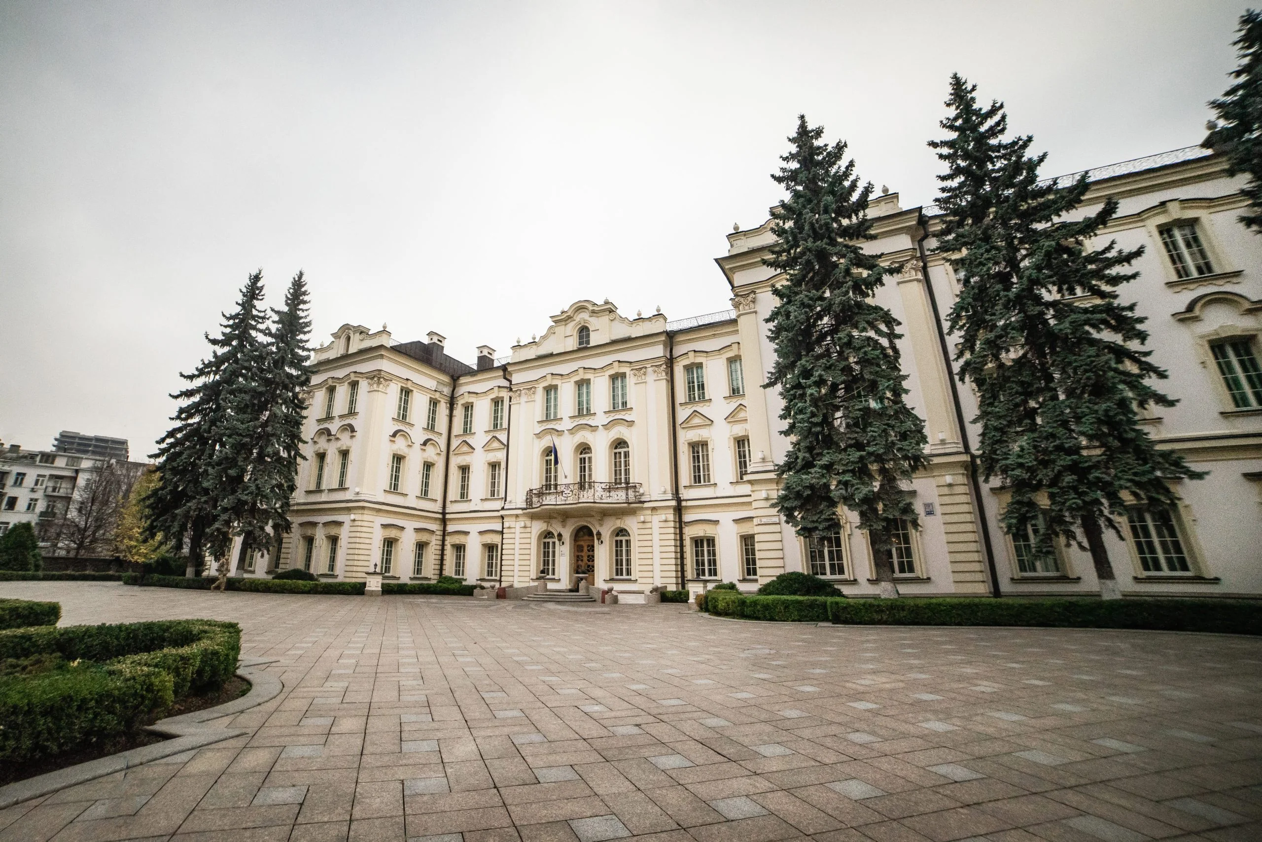 Білий класичний палац з колонами та українським прапором серед ялин на брукованій площі