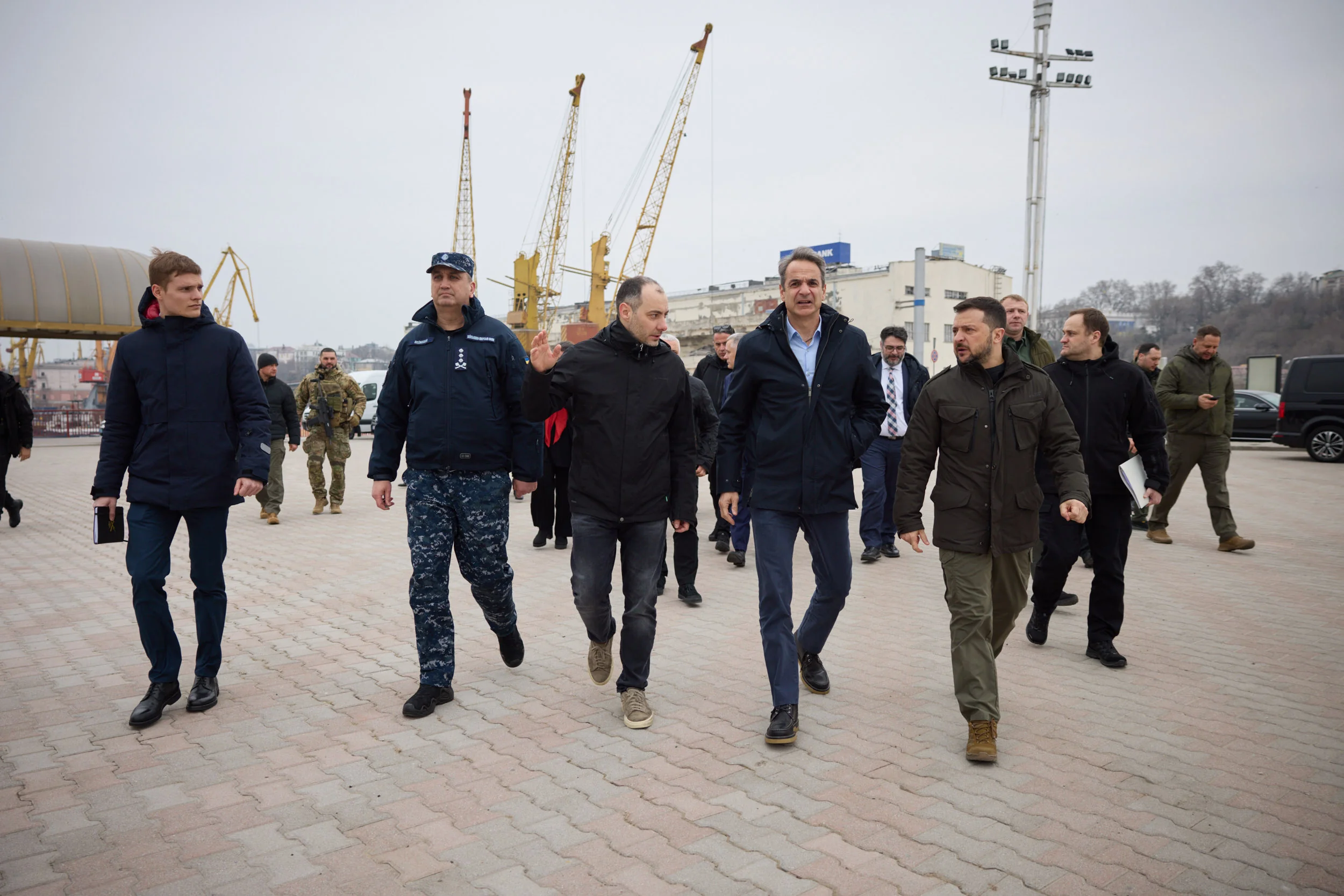 Група чоловіків у куртках йде портовою площею біля жовтих кранів