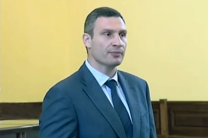 Віталій Кличко у темному костюмі стоїть на тлі жовтої стіни